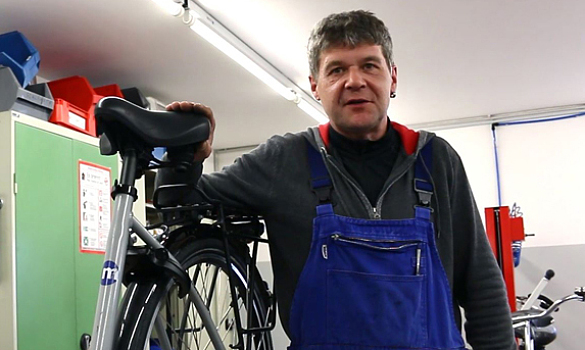 Ein Mann steht in einer Fahrradwerkstatt im Blaumann neben einem Rad.